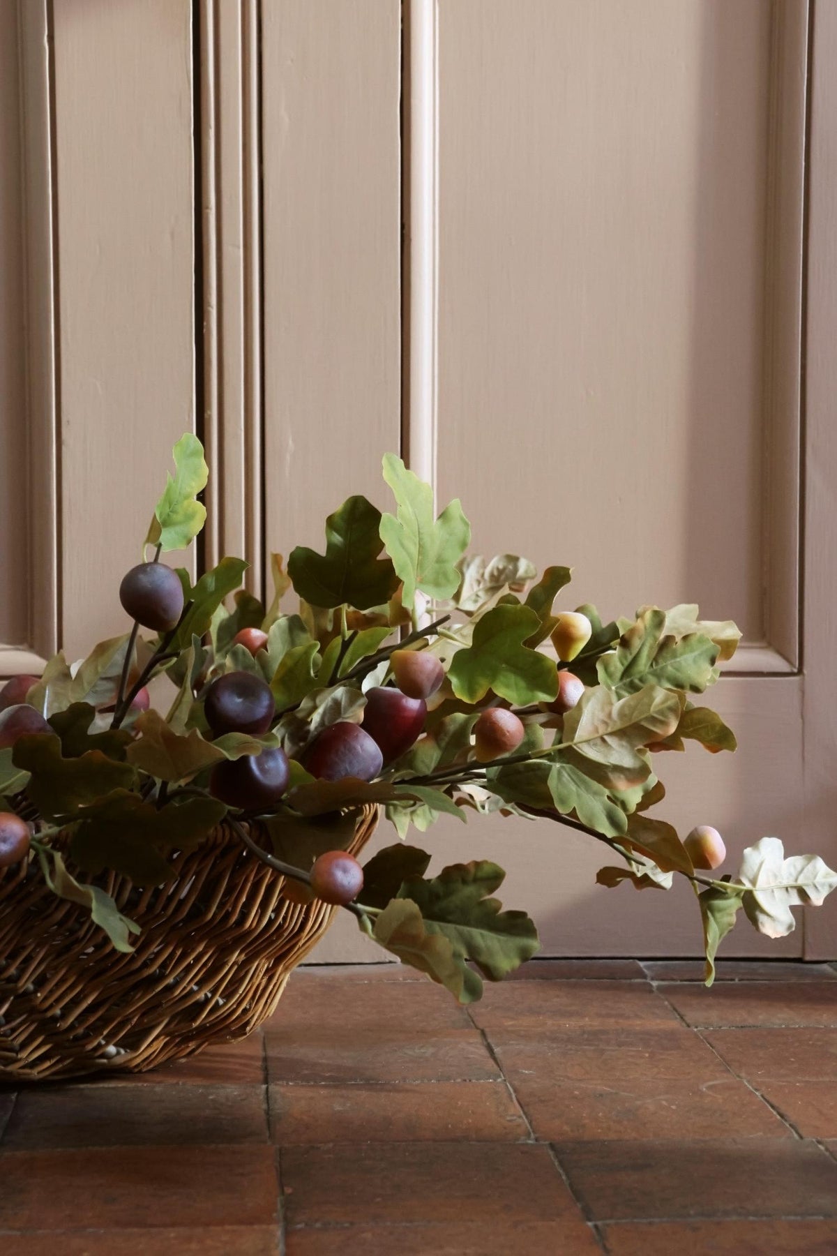 A wicker basket filled with faux oak leaves and small purple-brown figs, placed on a terracotta tile floor in front of a beige paneled wall.