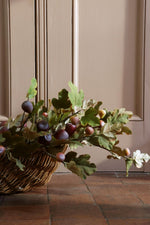 A wicker basket filled with faux oak leaves and small purple-brown figs, placed on a terracotta tile floor in front of a beige paneled wall.