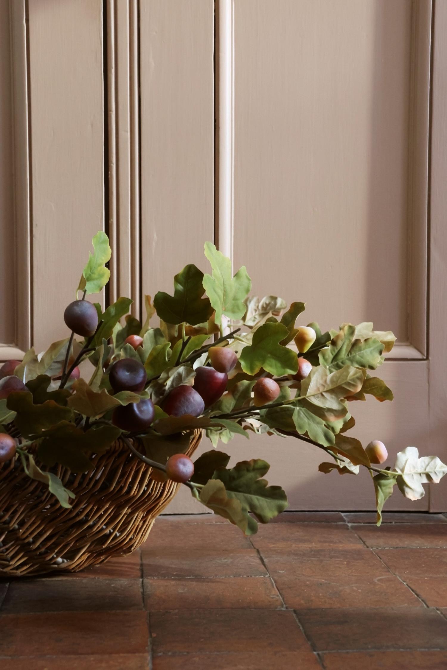 A wicker basket filled with faux oak leaves and small purple-brown figs, placed on a terracotta tile floor in front of a beige paneled wall.