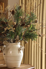 A rustic white ceramic vase filled with a natural pine branch arrangement featuring green needles, pinecones, and dried beige grasses, displayed on a wooden side table in a warm, neutral-toned room with beige curtains and wooden shelving.