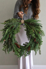 Person holding a large artificial foliage wreath with pinecones against a neutral background