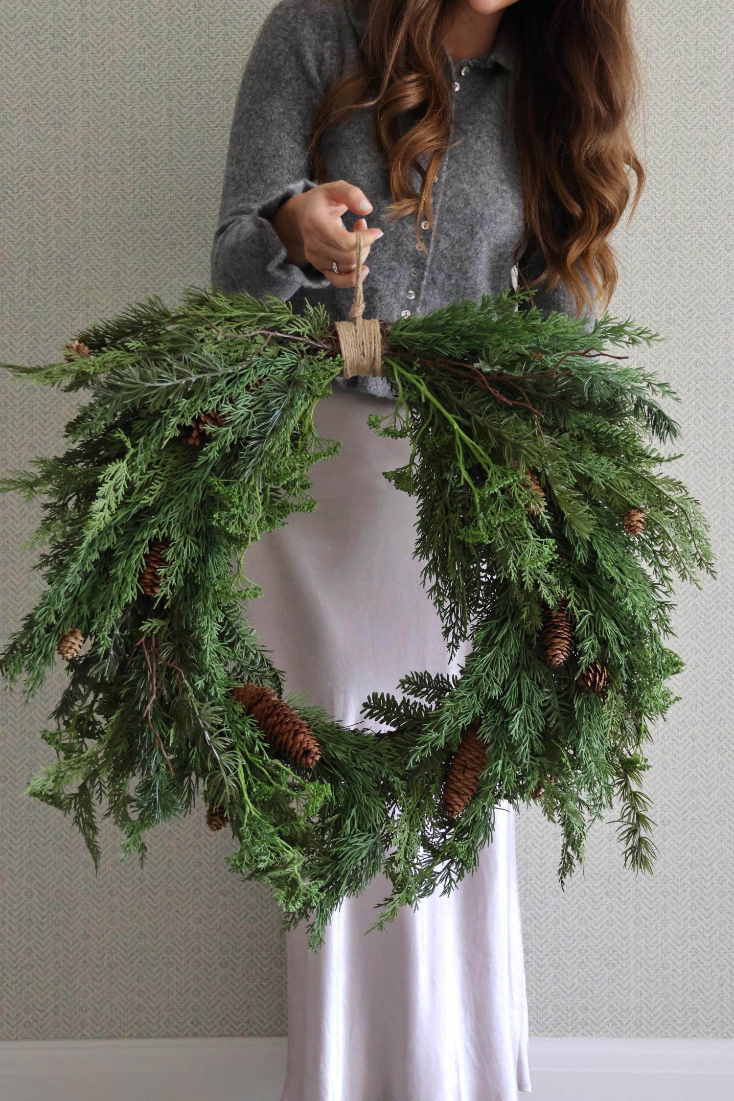 Person holding a large artificial foliage wreath with pinecones against a neutral background