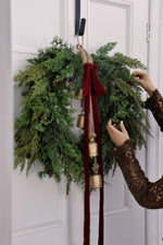 large artificial green foliage wreath with brass bells and a long red bow on white door 