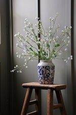 blue floral pattern Vase with faux white forget me not flowers on a wooden stool against a gray wall