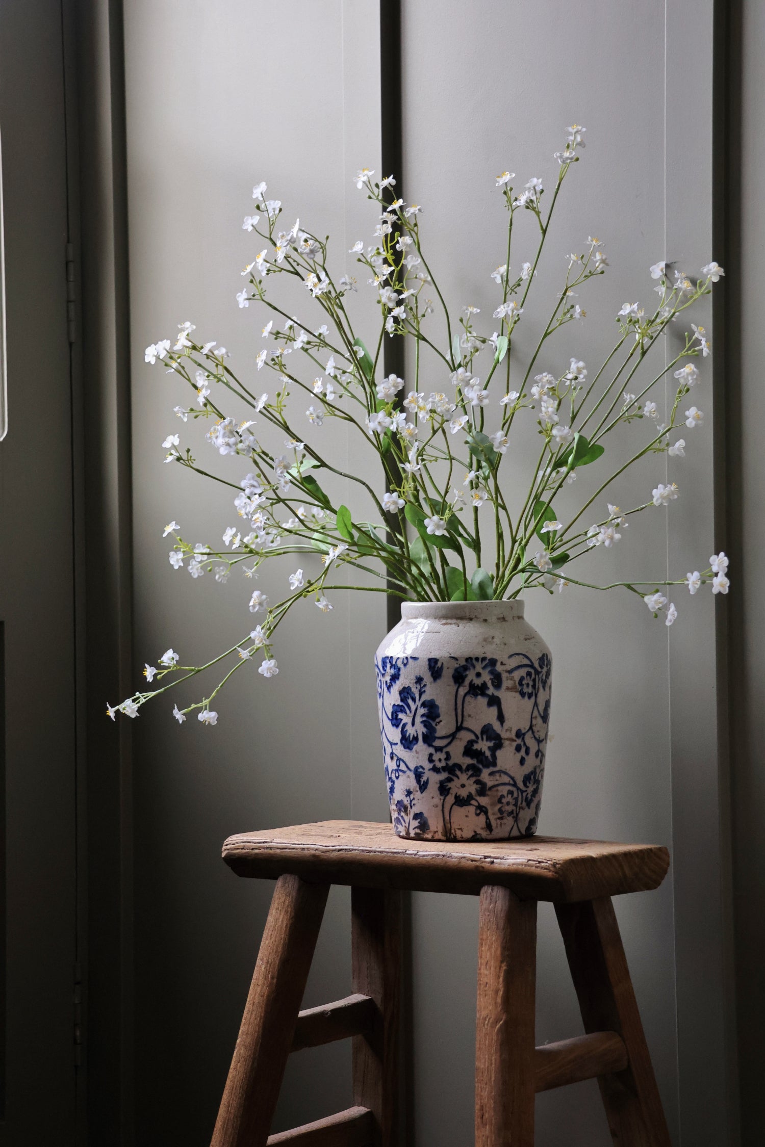 blue floral pattern Vase with faux white forget me not flowers on a wooden stool against a gray wall