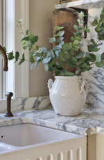 White vase with green eucalyptus on a marble countertop next to a kitchen sink.