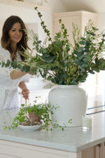 large artificial arrangement of green foliage in white vase on a kitchen island with women arranging behind