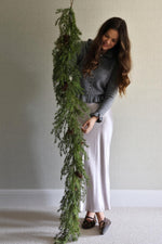 Woman holding a long artificial foliage garland against a plain wall.