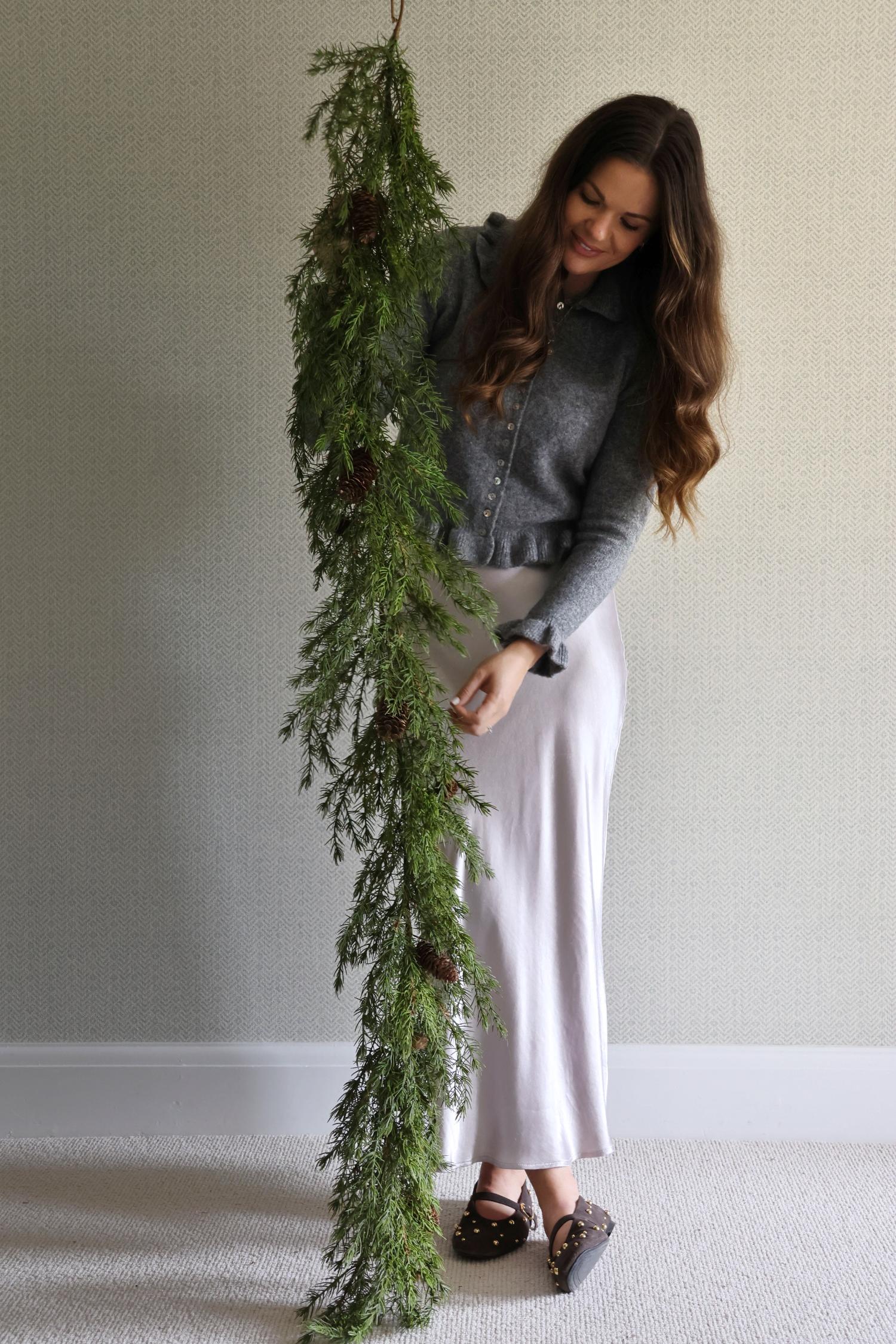 Woman holding a long artificial foliage garland against a plain wall.