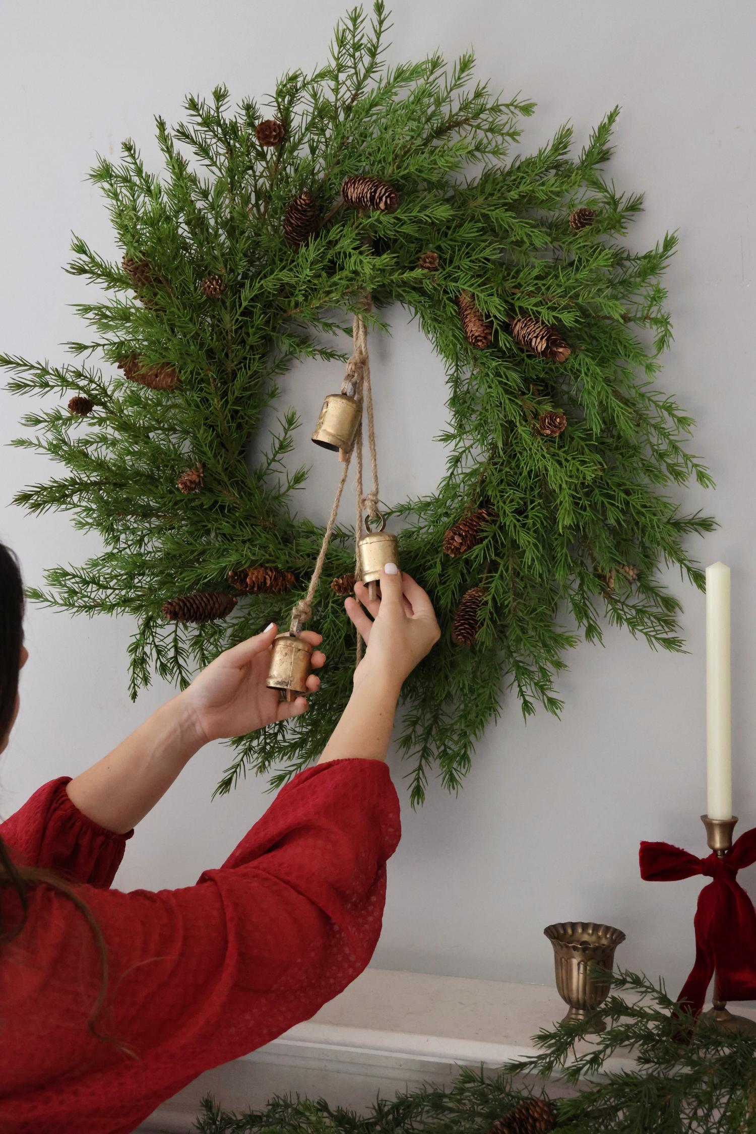 Person adjusting an artificial festive wreath with brass hanging cowbells on a wall.