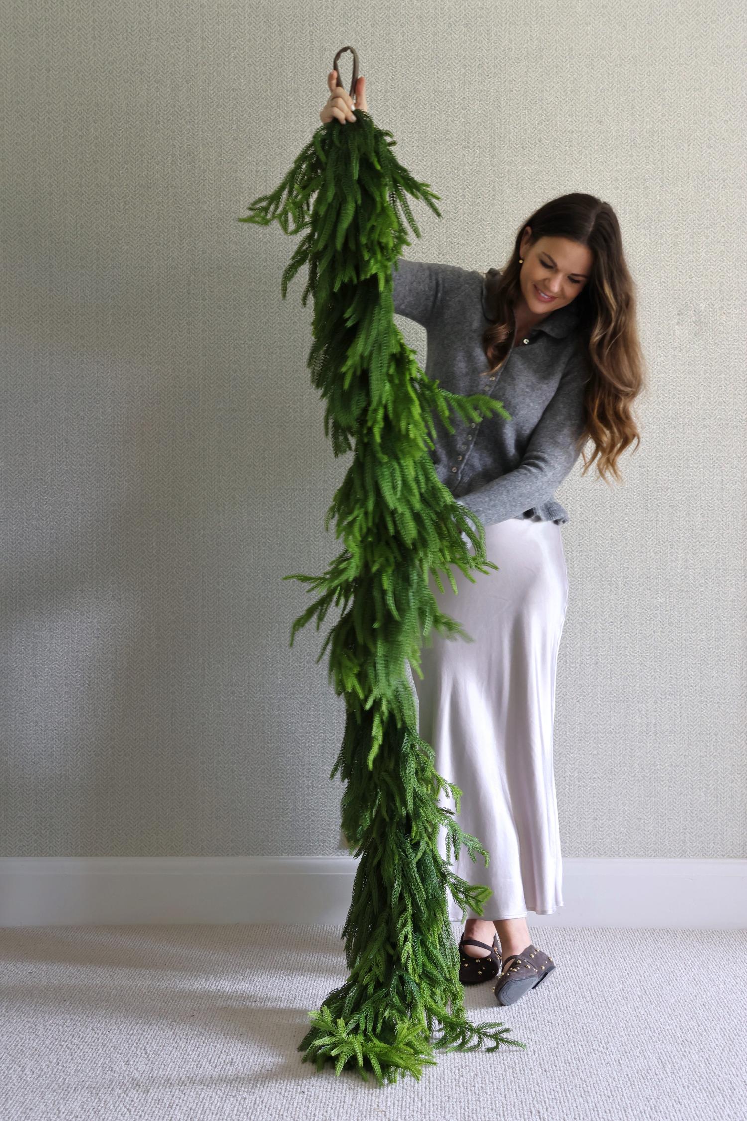 Woman holding a large artificial norfolk pine garland against a plain wall.