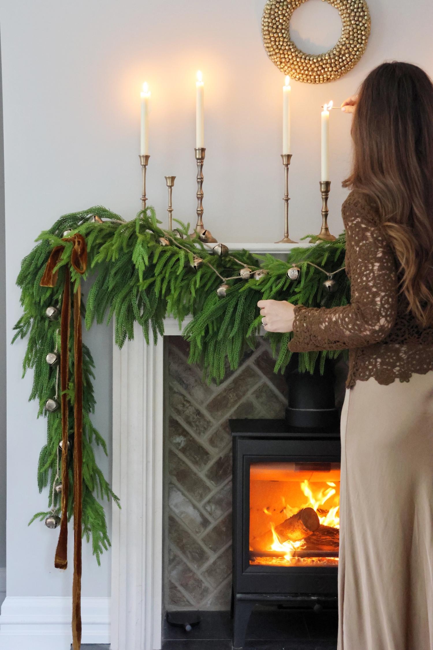 Woman by a fireplace with artificial norfolk pine garland, bells, ribbon and candles