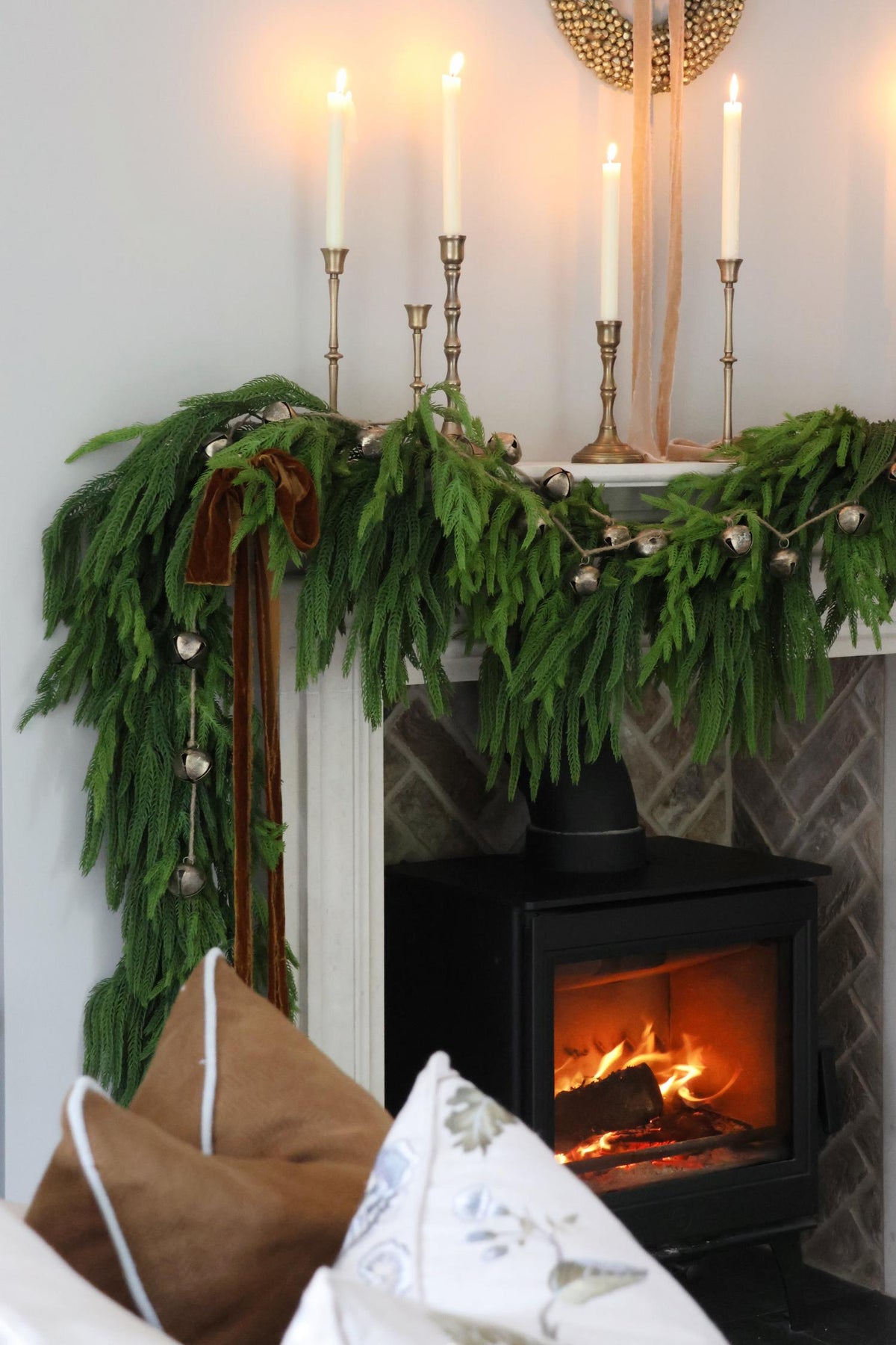 Fireplace with artificial norfolk pine garland and candles on a mantel, surrounded by cozy pillows.