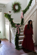 Woman in a red dress standing on a staircase decorated with artificial norfolk pine garlands, a large faux wreath and bells and ribbons.