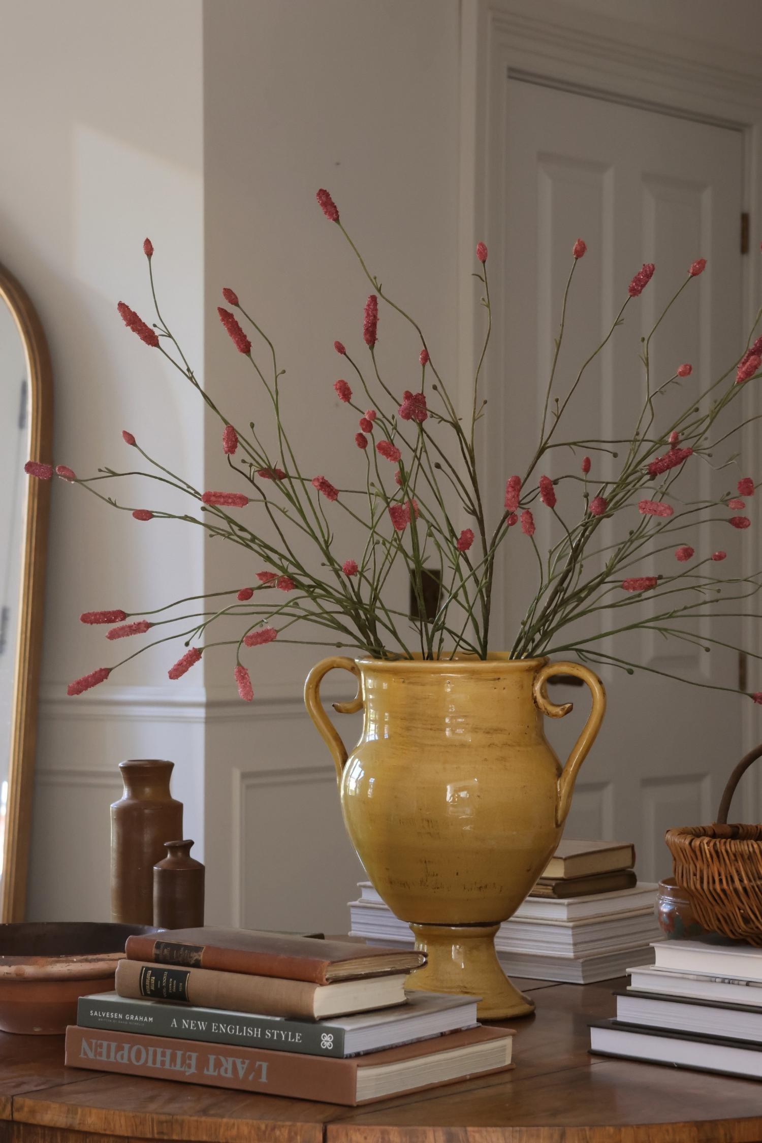 A yellow ceramic urn-shaped vase filled with artificial burnet pink wildflowers, styled on a wooden table with books in a bright room.