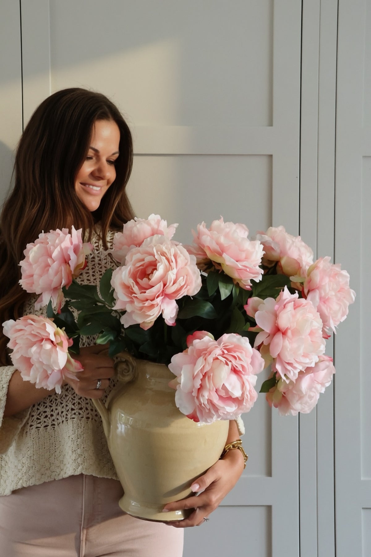 Woman holding a large cream jug filled with faux pink peony flowers against a light gray wall.