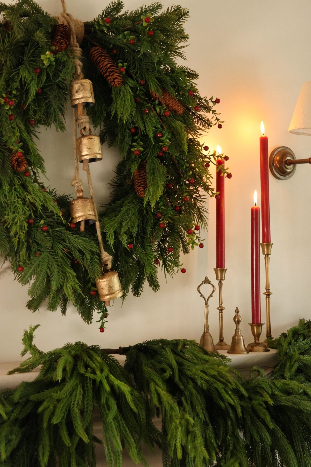 a large artificial christmas wreath with rustice hanging cowbells, red berries and pinecones above a mantel with brass candlesticks and lit red candles and a norfolk pine garland 