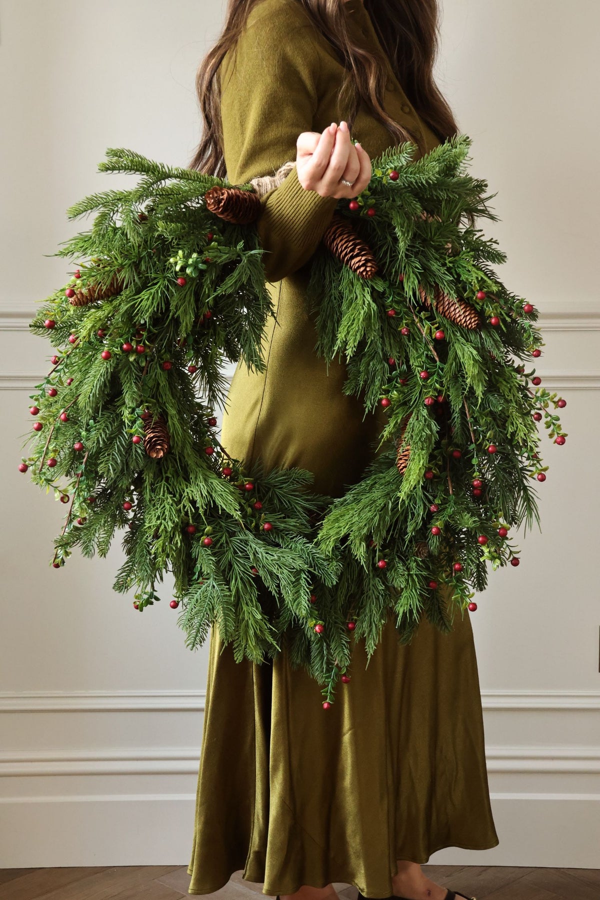 women in long green dress holding a large artificial wreath with green foliage, red berries, and pinecones 