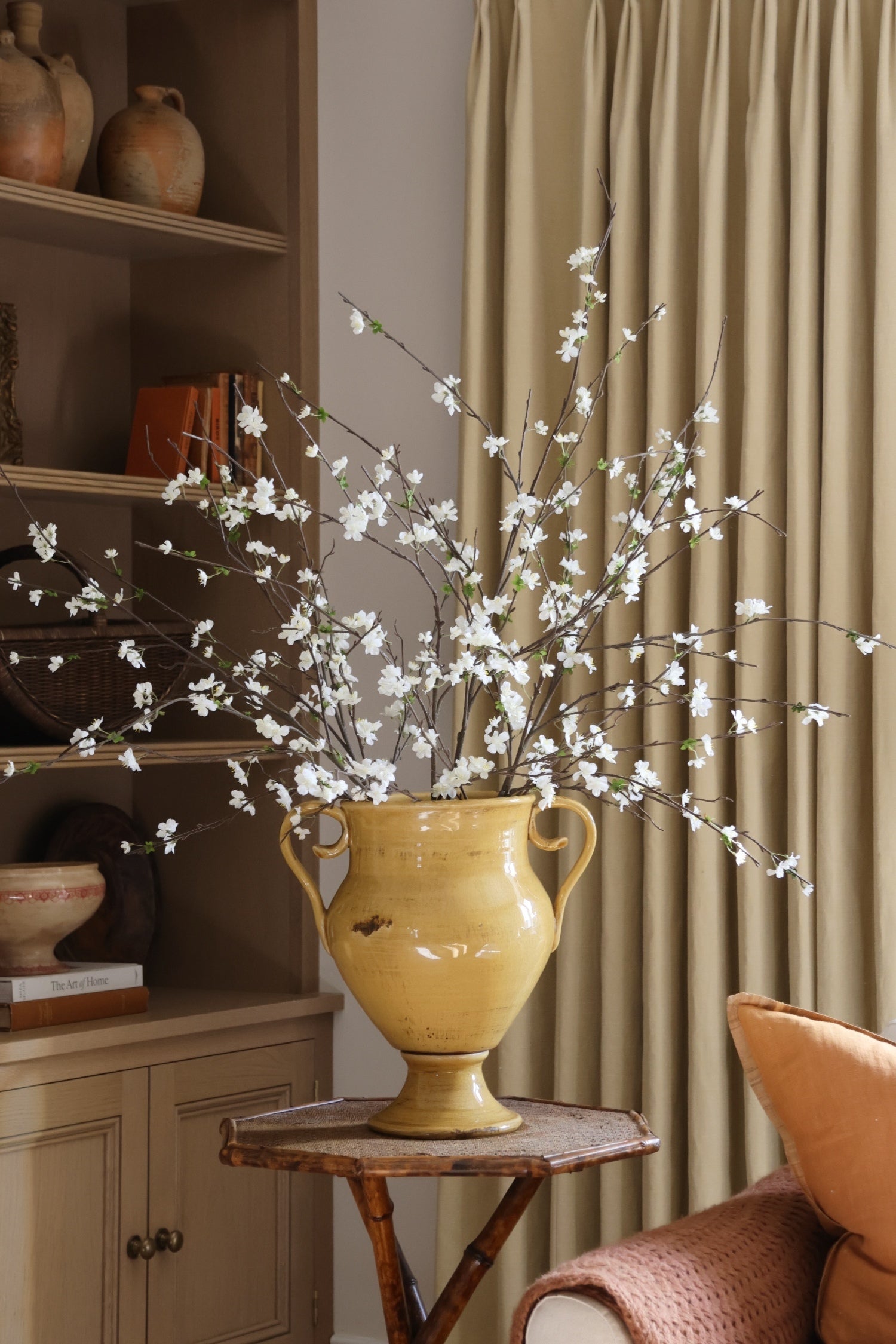 Yellow urn shaped vase with faux white sakura branches on a small wooden table against a beige curtain.