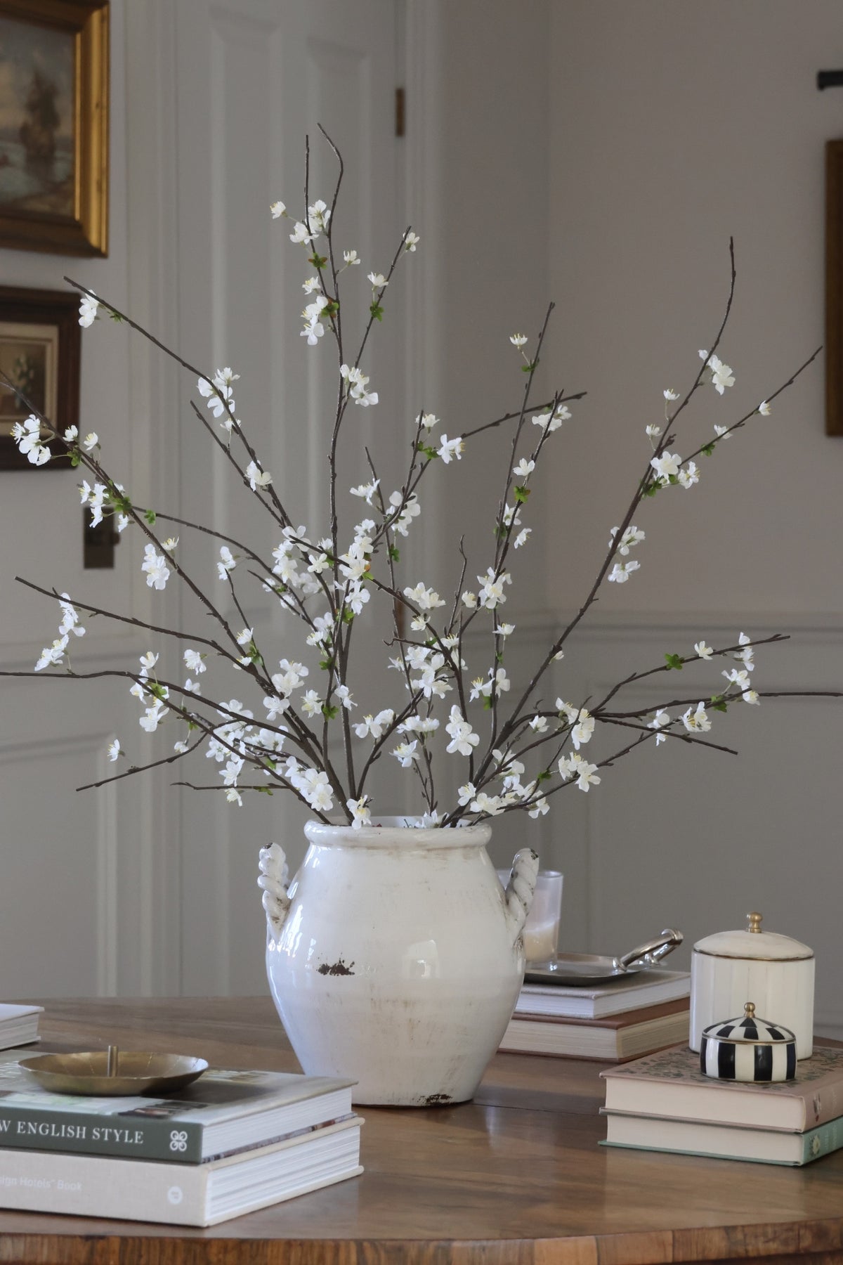 White vase with artificial blossom branches on a wooden surface with books and decorative items.
