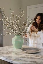 Woman arranging artificial sakura blossom branches in a green vase on a marble countertop.
