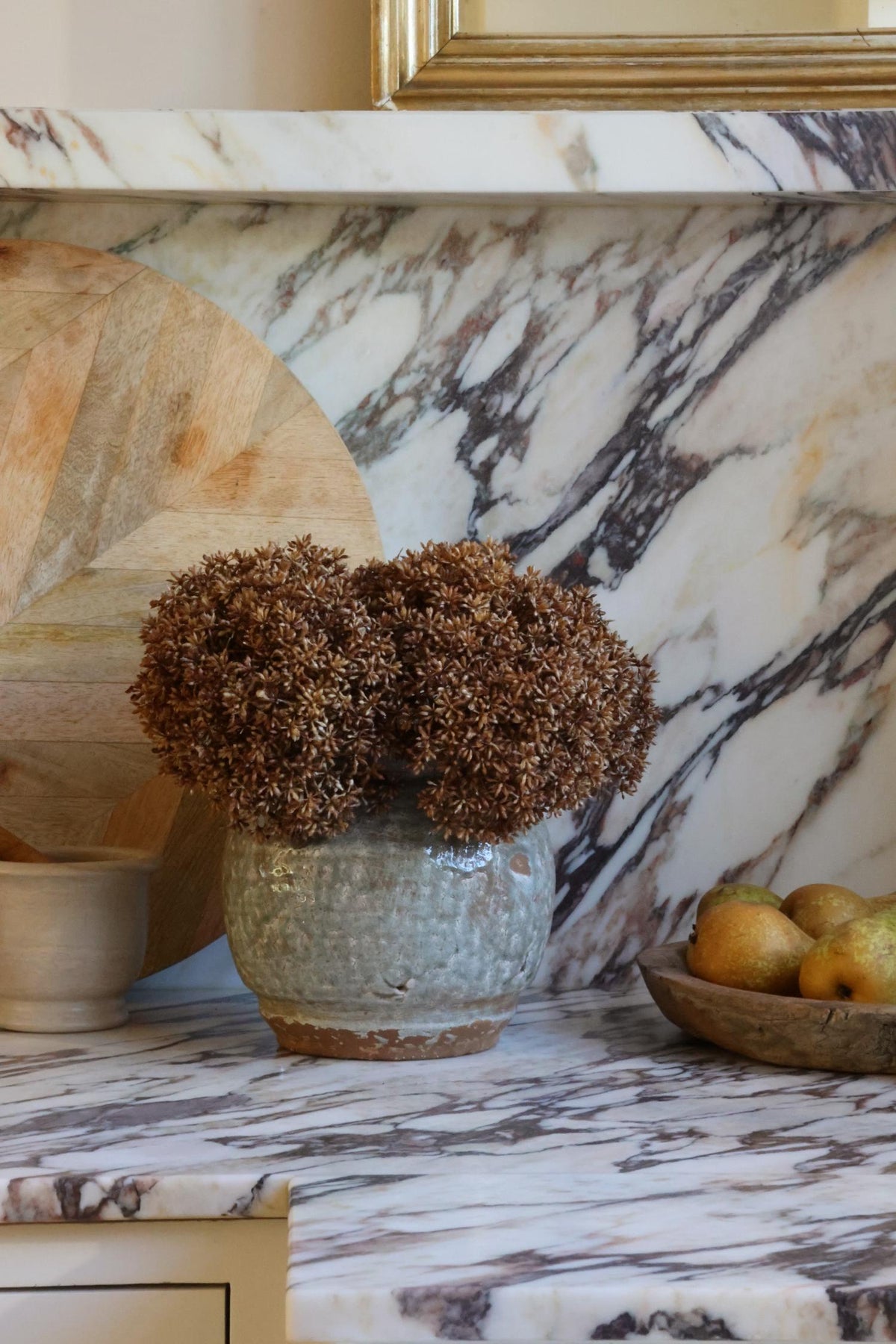 A rustic ceramic pot filled with artificial dried brown flowers, styled on a white and gray marble countertop with a matching marble backsplash, a wooden cutting board, and a bowl of pears in the background.