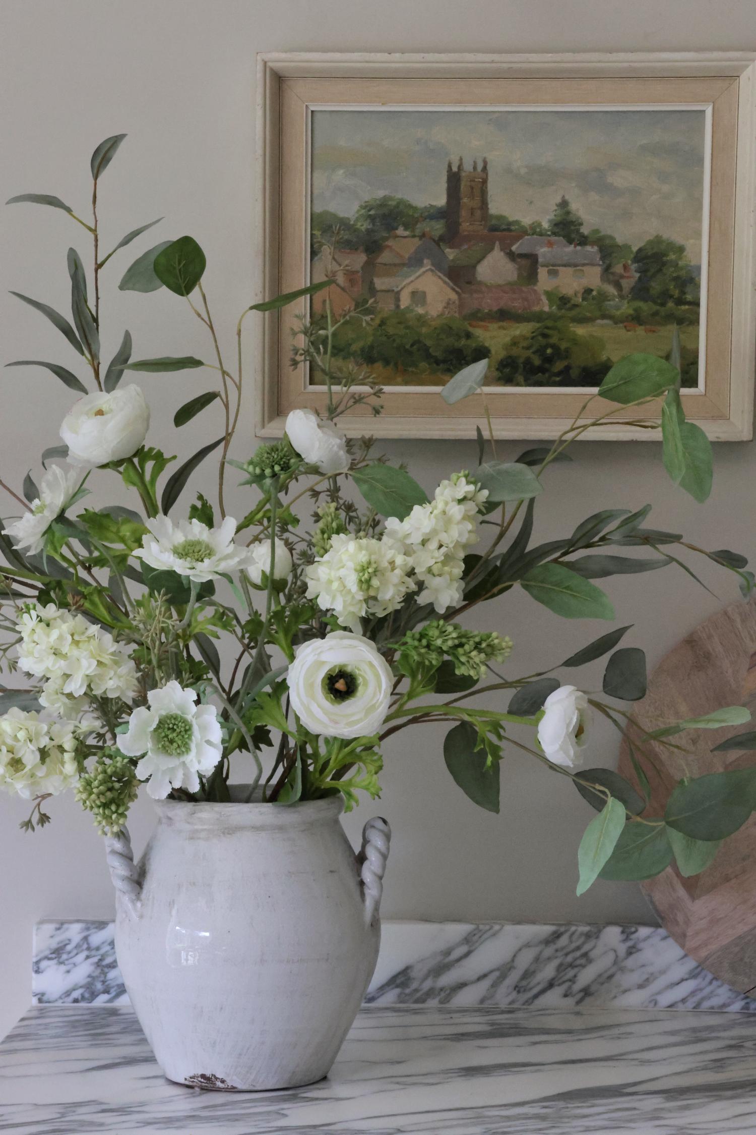 Floral arrangement in a white vase on a marble surface with a framed painting in the background.