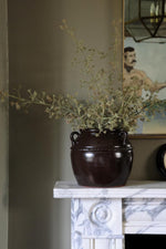 A faux seeded eucalyptus stem in a brown vase placed on a white surface, with a painting reflecting in the mirror.