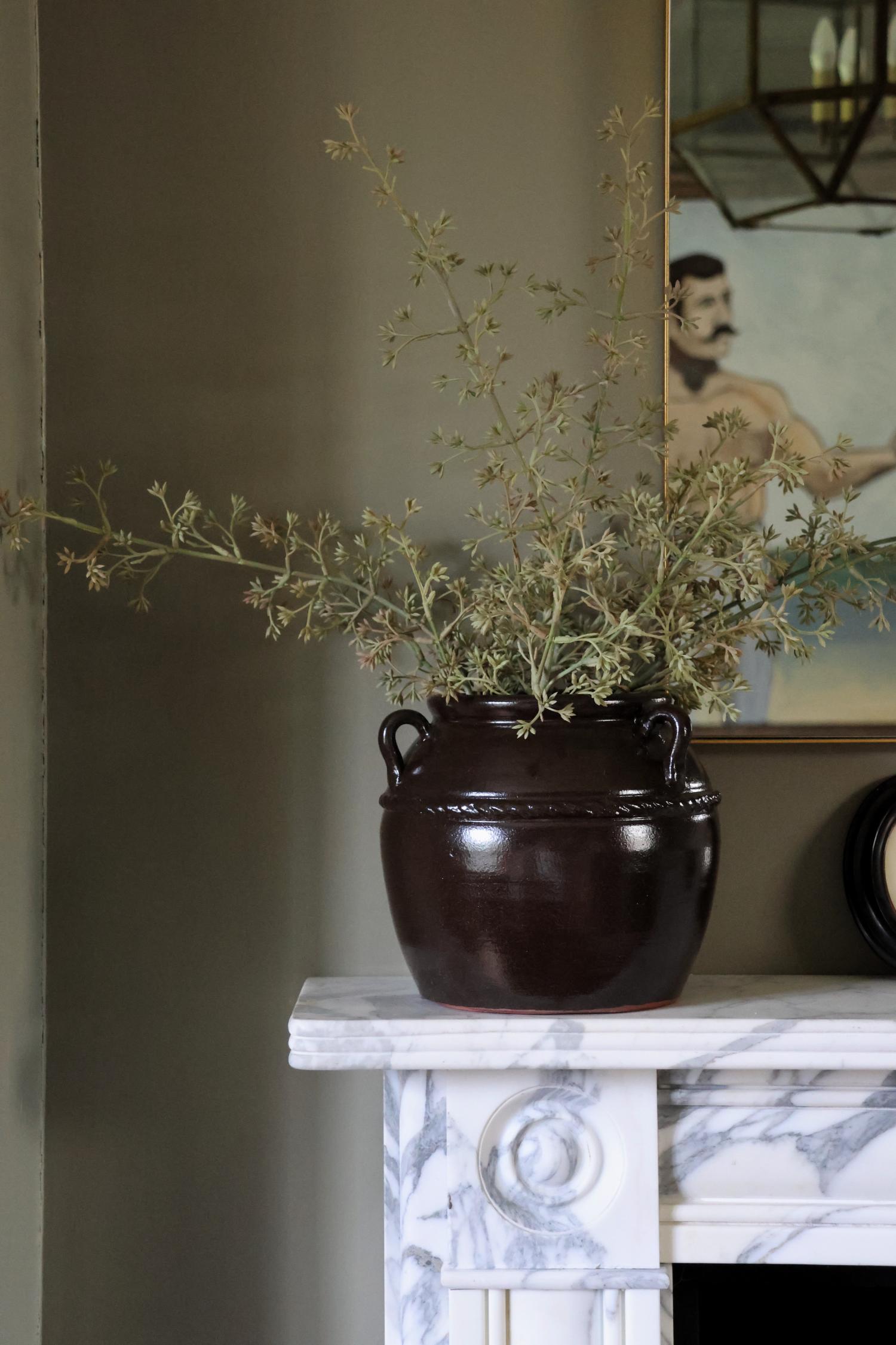 A faux seeded eucalyptus stem in a brown vase placed on a white surface, with a painting reflecting in the mirror.