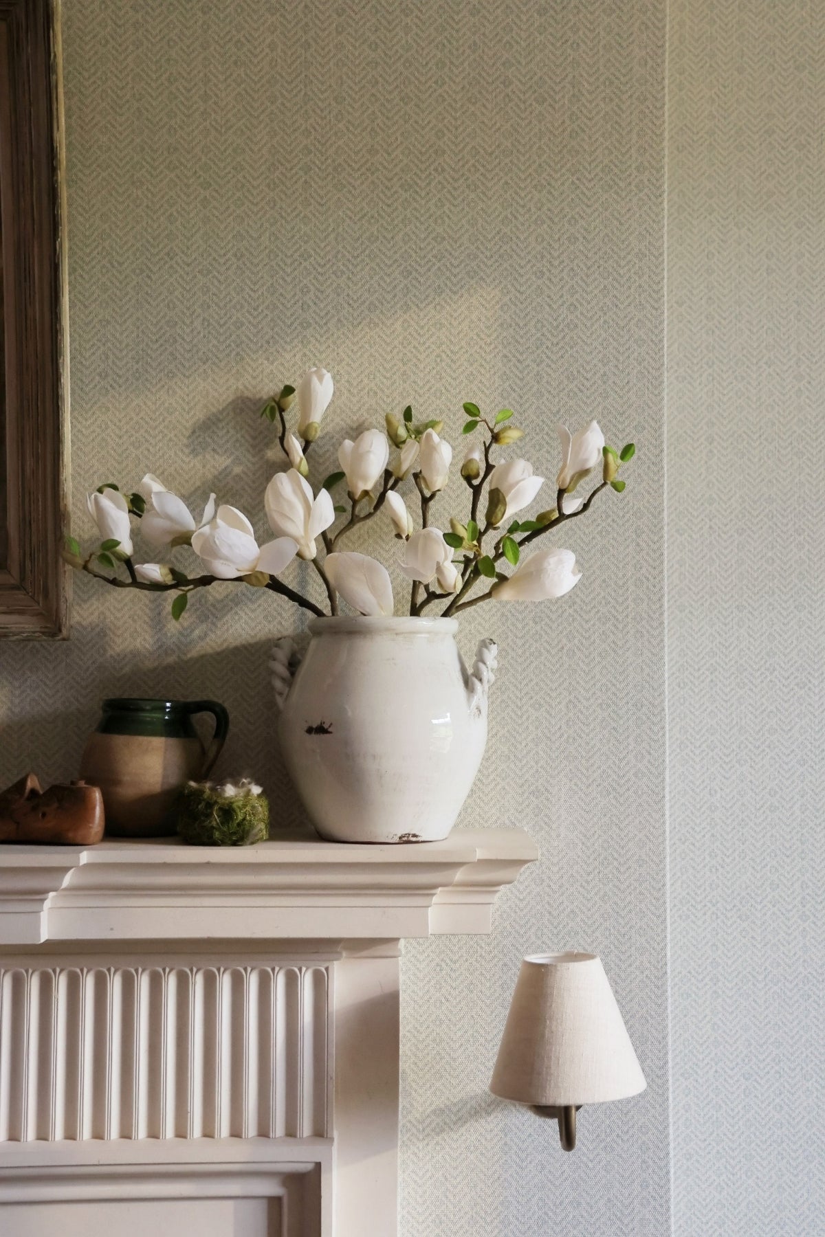 White vase with flowers on a fireplace mantle against a light gray wall.