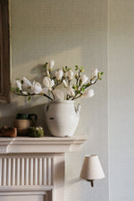 White vase with flowers on a fireplace mantle against a light gray wall.
