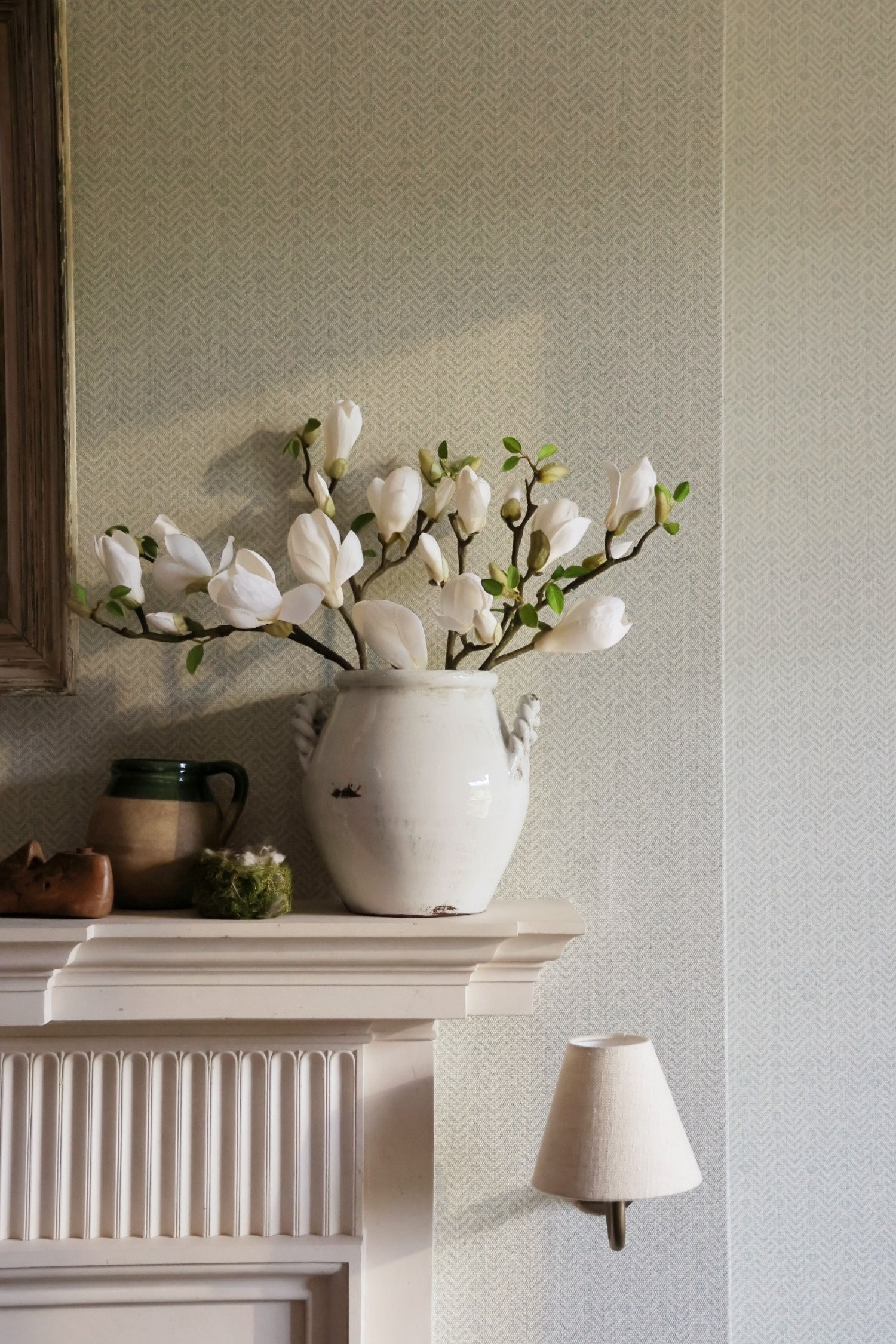 White vase with flowers on a fireplace mantle against a light gray wall.