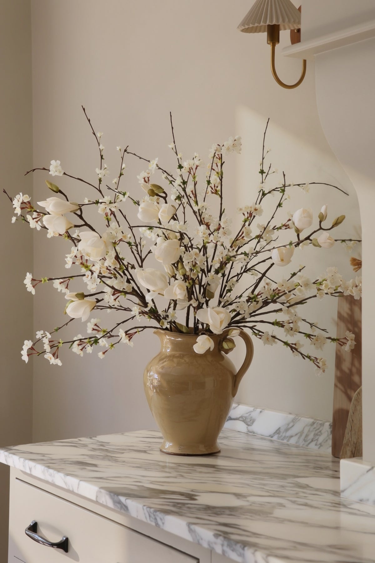 Beige ceramic cavendish jug with white artificial spring branches arrangement on a marble surface