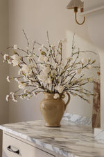 Beige ceramic cavendish jug with white artificial spring branches arrangement on a marble surface