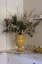 A large mustard yellow ceramic urn vase filled with a winter faux floral arrangement of green pine branches, white berries, and brown twigs, displayed on a white kitchen counter.