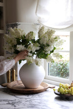 A person arranging artificial white lilac stems in a white vase, with the stems appearing realistic and touchable.