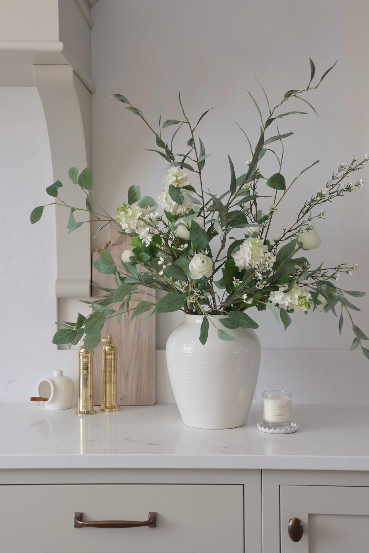 white vase in kitchen setting wiht green foliage and white artificial flowers