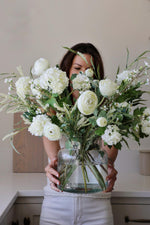 white and green artificial floral arrangement in a glass vase held by a woman