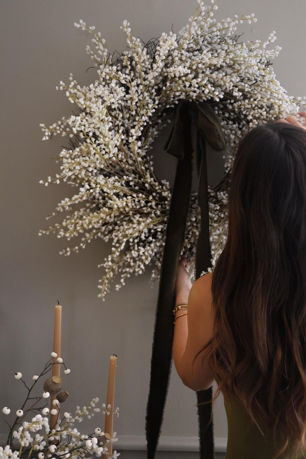 women arranging large artificial white willow wreath with dark green ribbon on a plain wall 