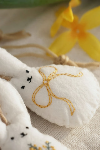 White fabric bunny with yellow bow embroidery on a textured surface with yellow flowers in the background