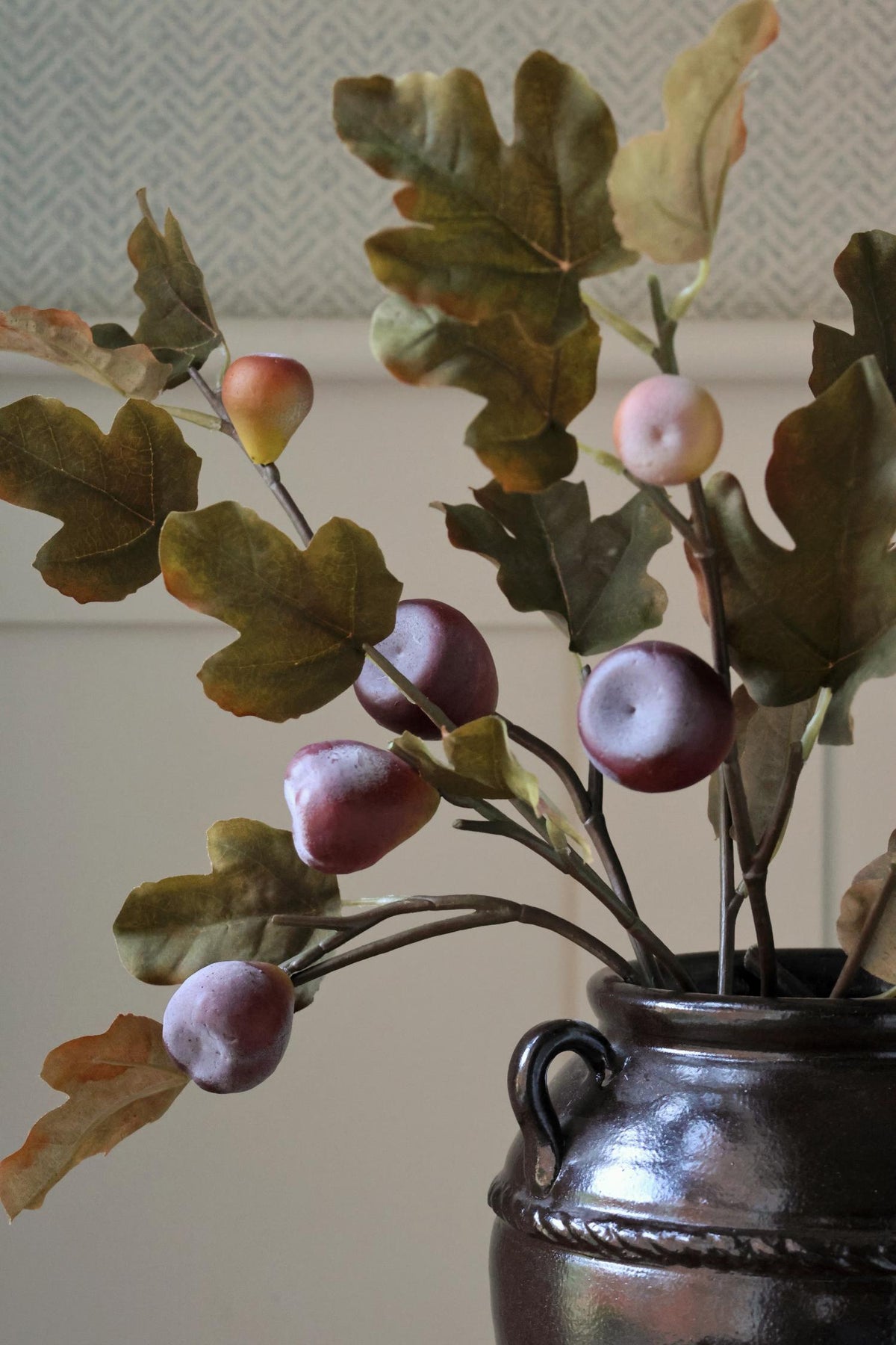 Artificial autumn fig branch arrangement with realistic brown leaves and pinkish-purple acorns, displayed in a dark metallic ceramic pot against a neutral indoor background.