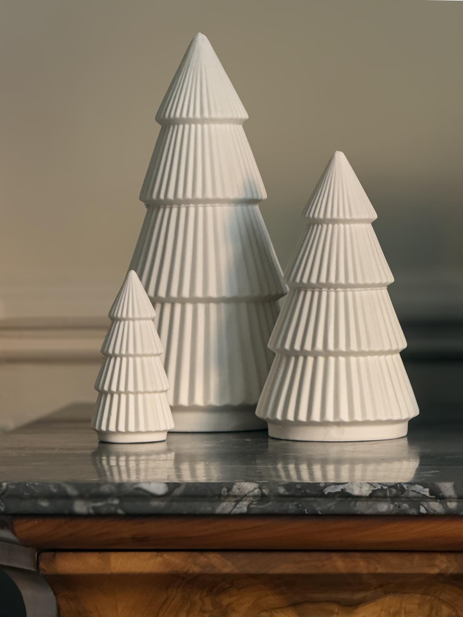 Three white ceramic Christmas tree figurines on a reflective surface with a neutral background