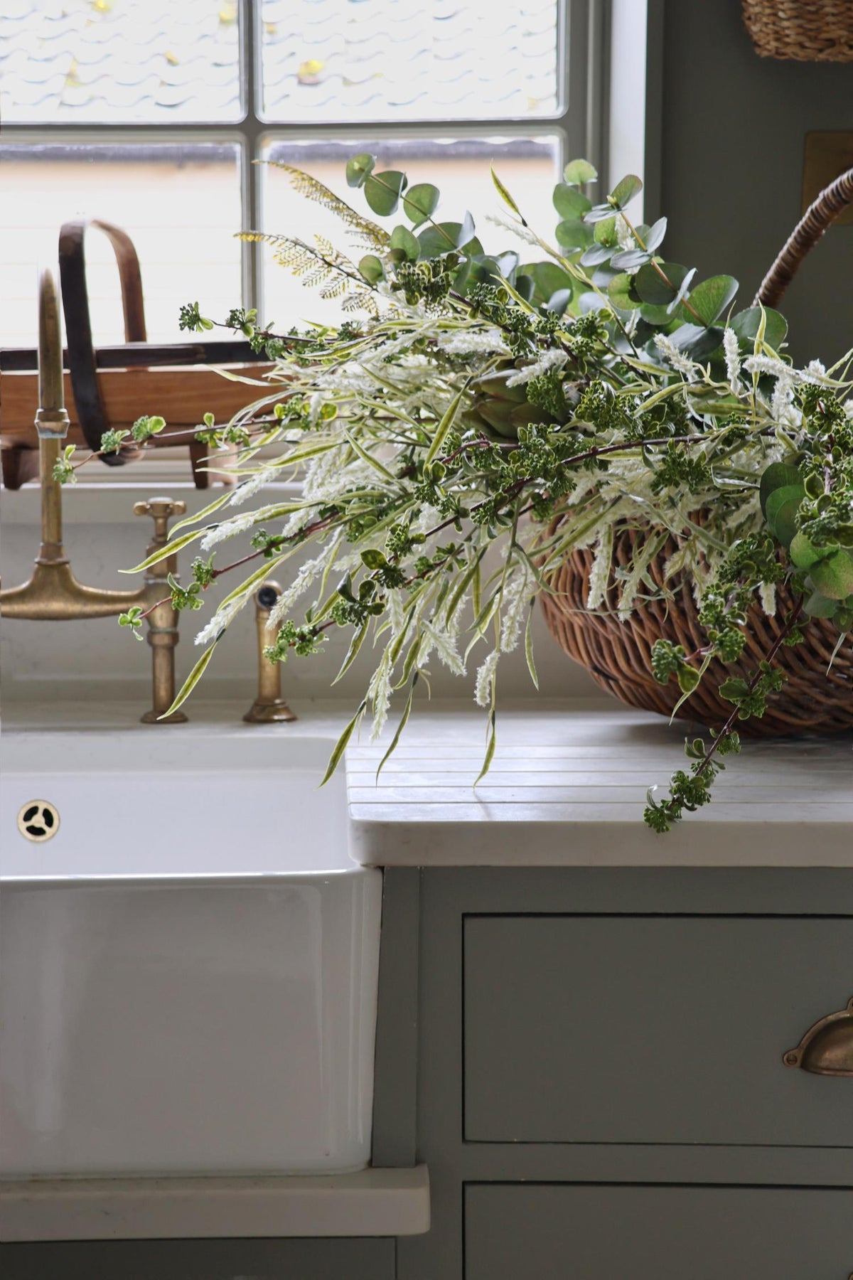 mixture of textured green foliage stems in a wicker basket on bootroom kitchen worktop and butler sink