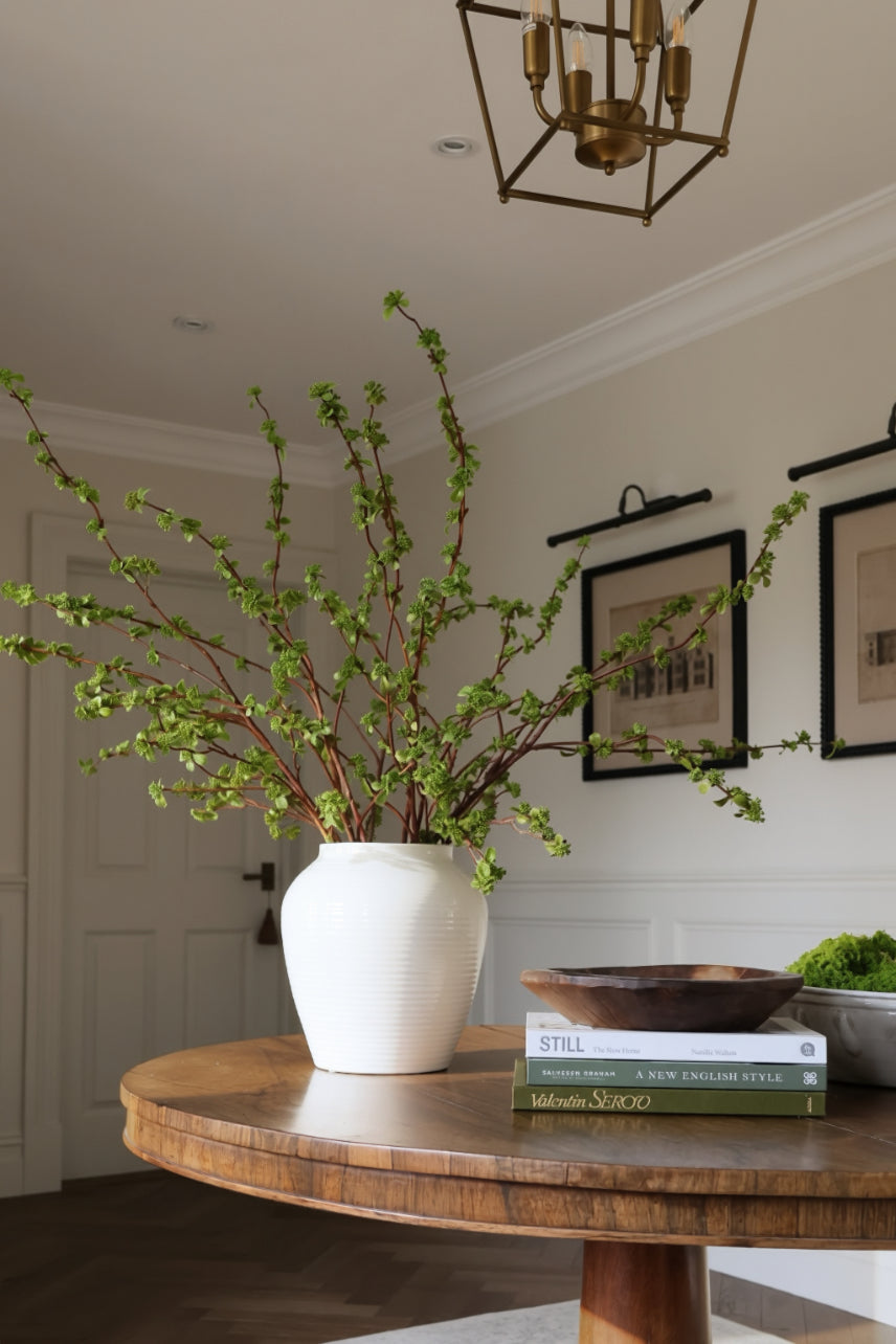 A large white vase filled with green berry branches, styled on a round wooden table in a bright, modern room with framed wall art and a brass chandelier.