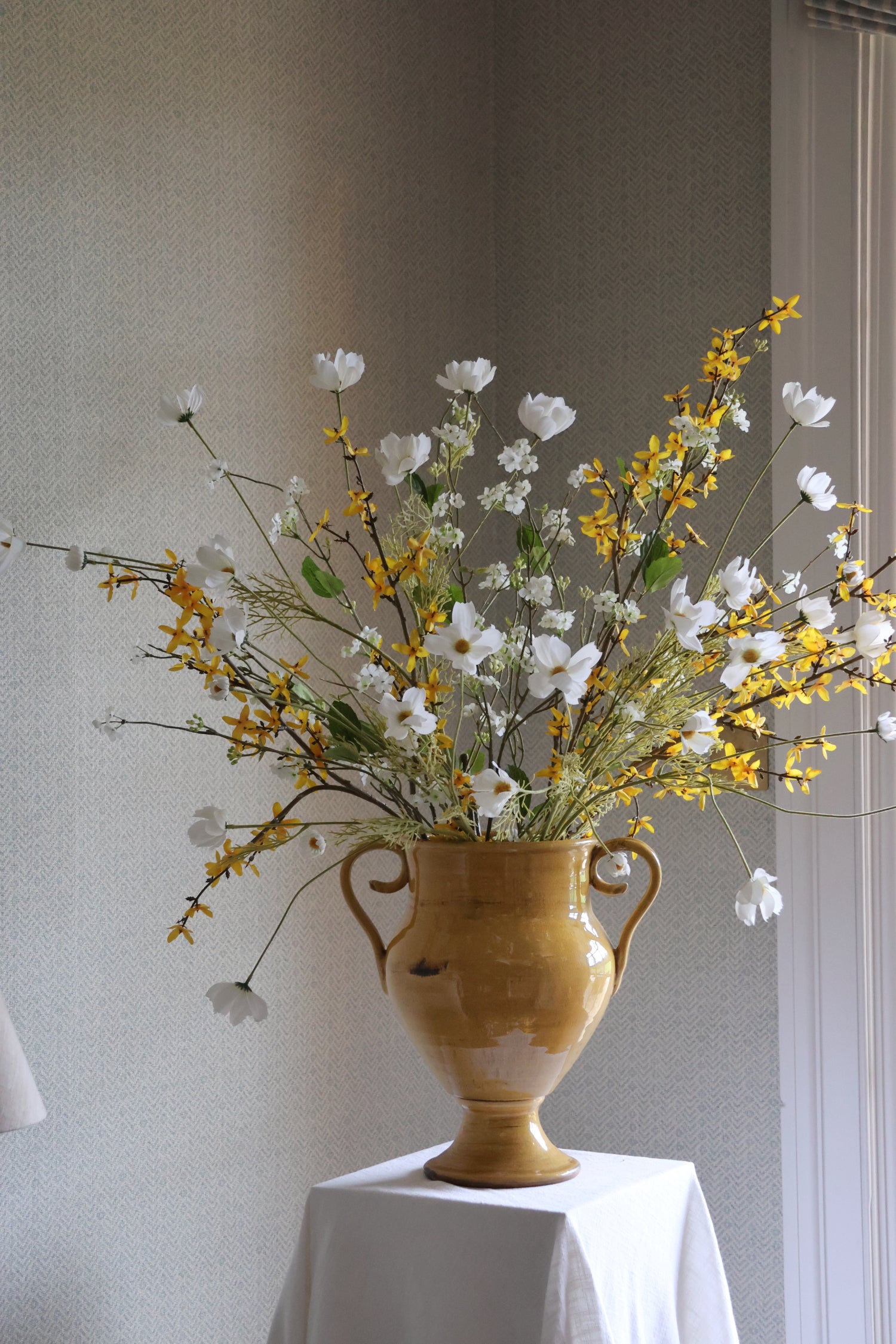 Yellow Vase with flowers on a white tablecloth against a neutral wall
