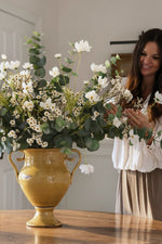 Woman arranging white and green flowers in a large yellow vase on a wooden table.