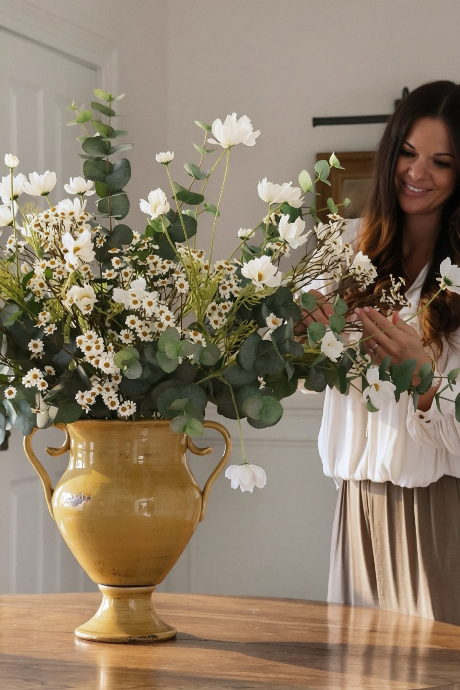 Woman arranging white and green flowers in a large yellow vase on a wooden table.