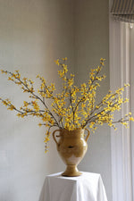 Yellow faux forsythia branches arrangement in a dark yellow vase on a white surface with a neutral background