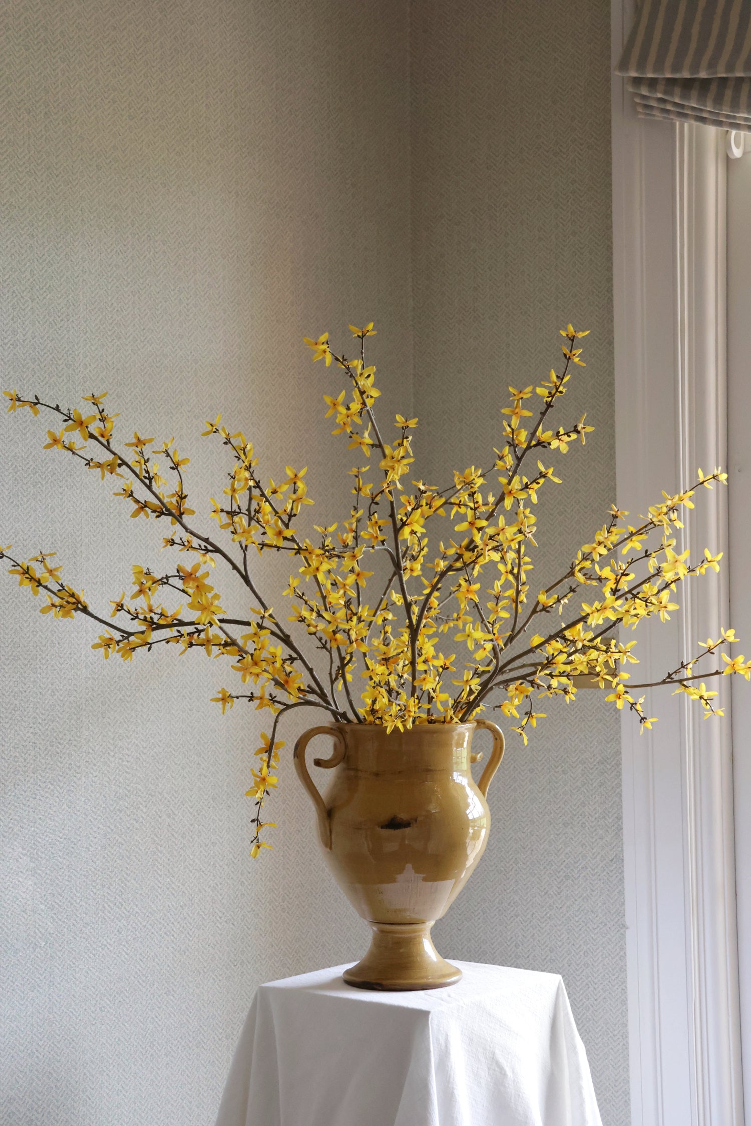 Yellow faux forsythia branches arrangement in a dark yellow vase on a white surface with a neutral background