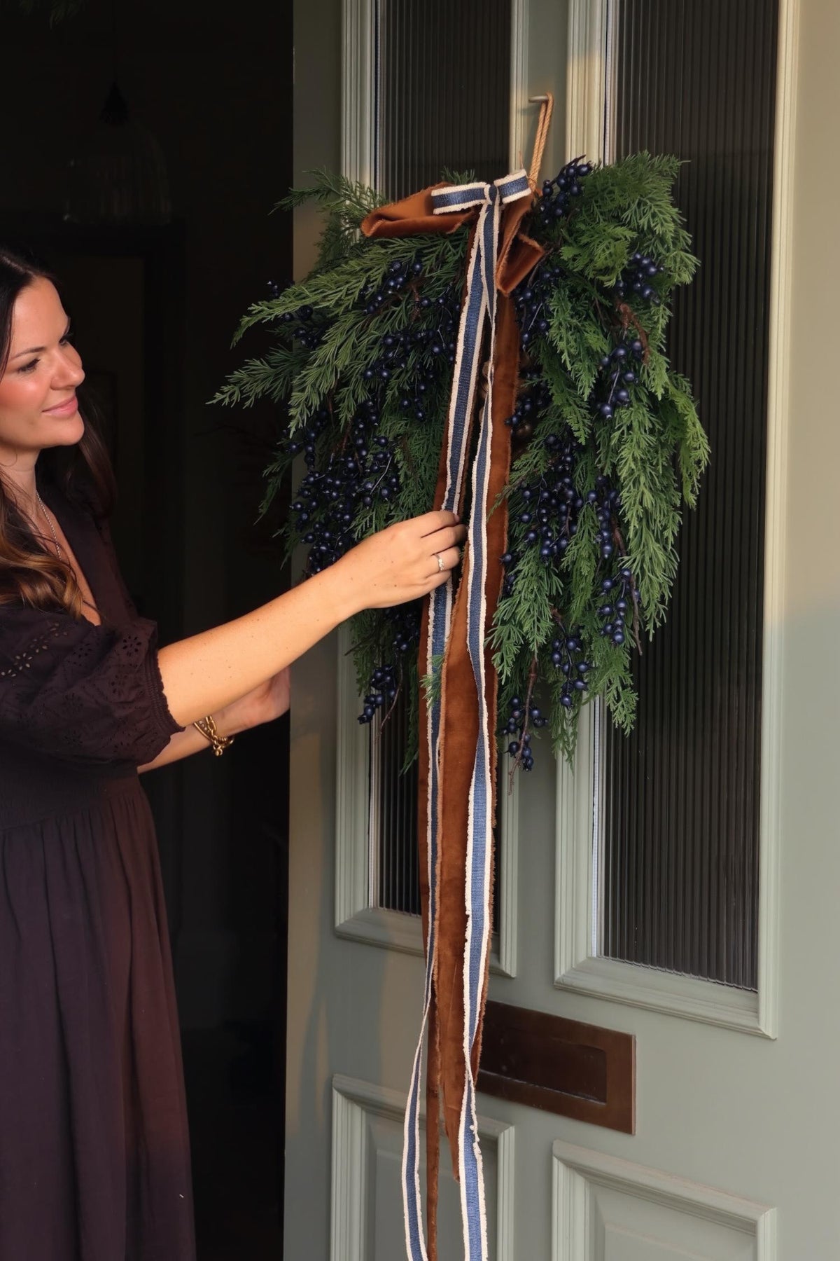 A woman in a dark plum dress is adjusting a large holiday wreath hanging on a white paneled door. The wreath is made of green evergreen branches, decorated with dark blue berries, and tied with a long brown ribbon with blue and white stripes.